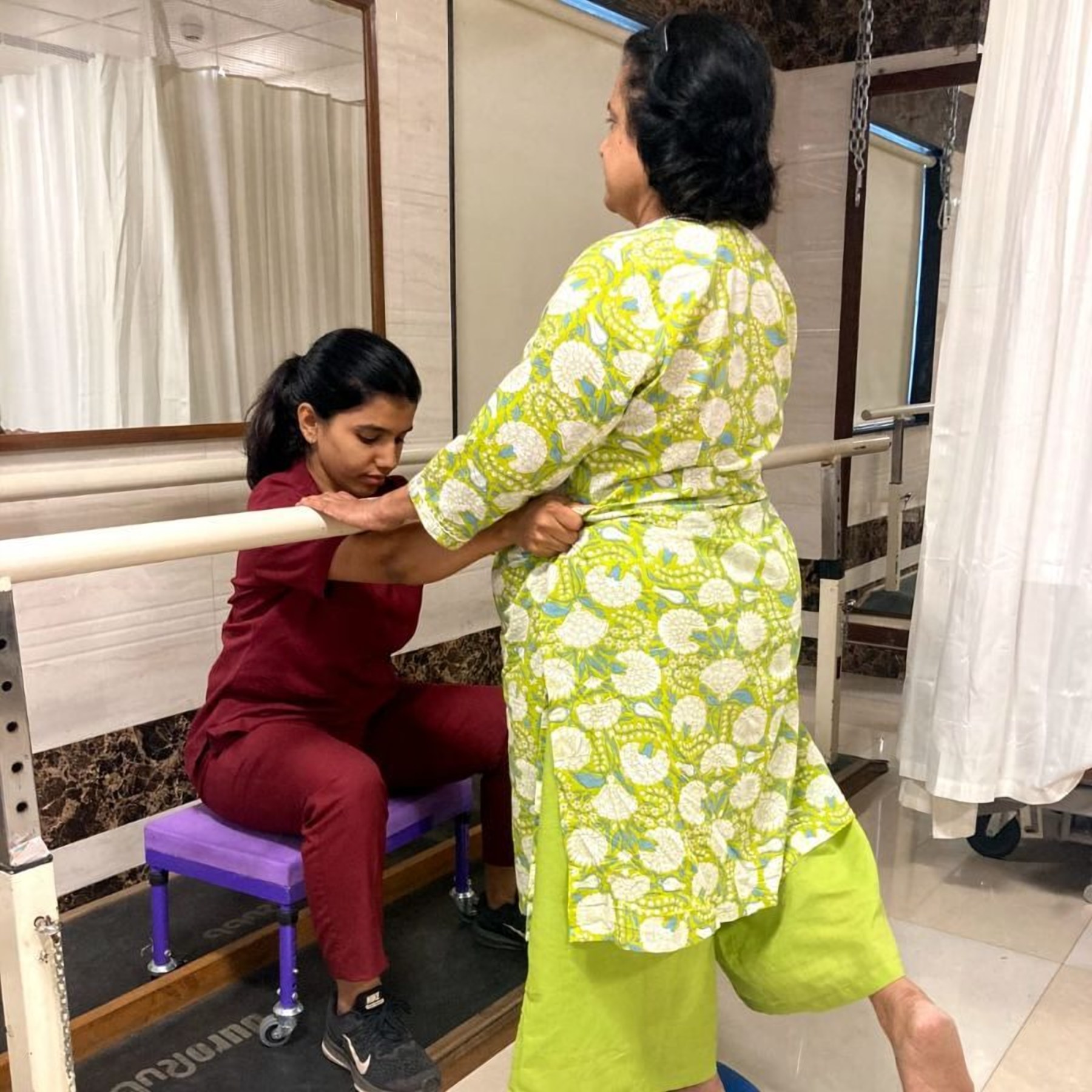 A Faith Physical Therapy therapist steadying an older patient walking the parallel bars in Thane